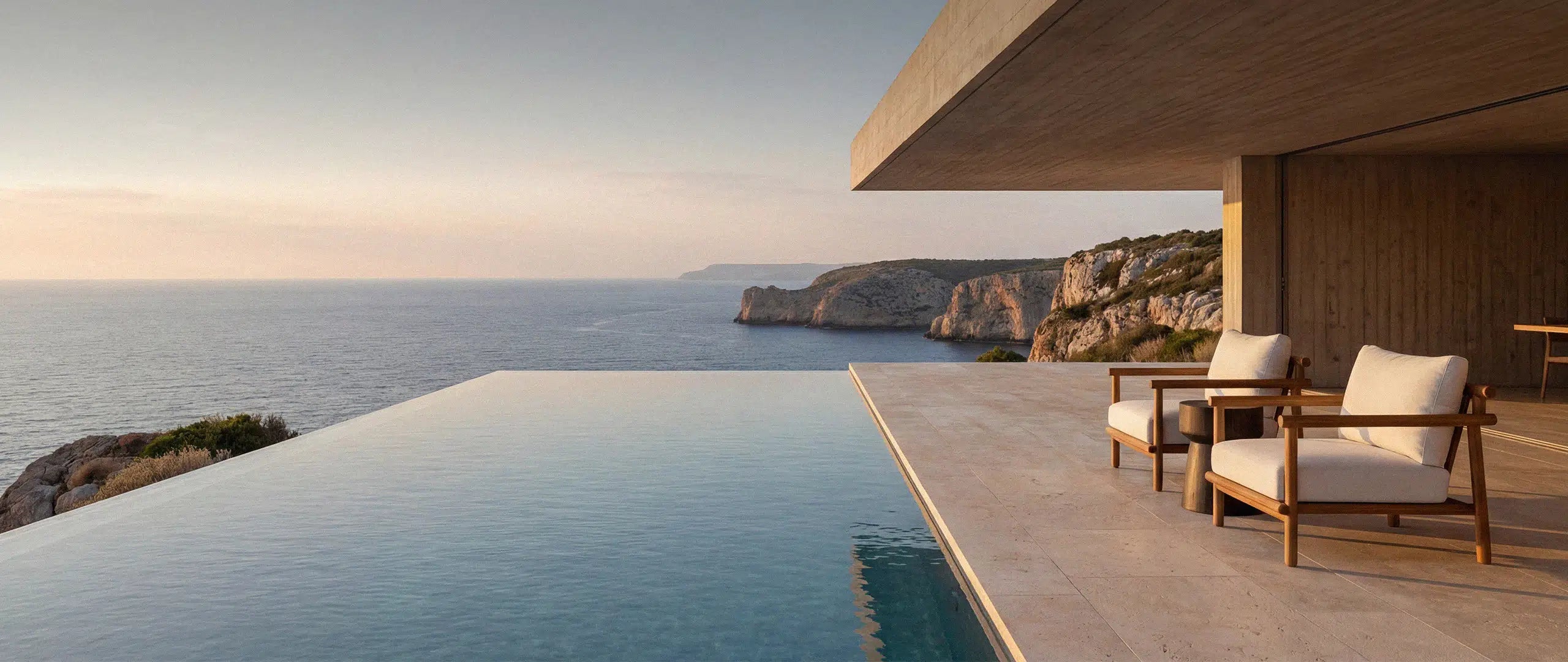 Zwei exklusive Menelamé Teakholz-Sessel mit weißen Polstern auf einer luxuriösen Villa-Terrasse mit Infinity-Pool. Die goldene Abendsonne über der Meeresküste unterstreicht das erstklassige Design unserer Outdoor-Kollektion.