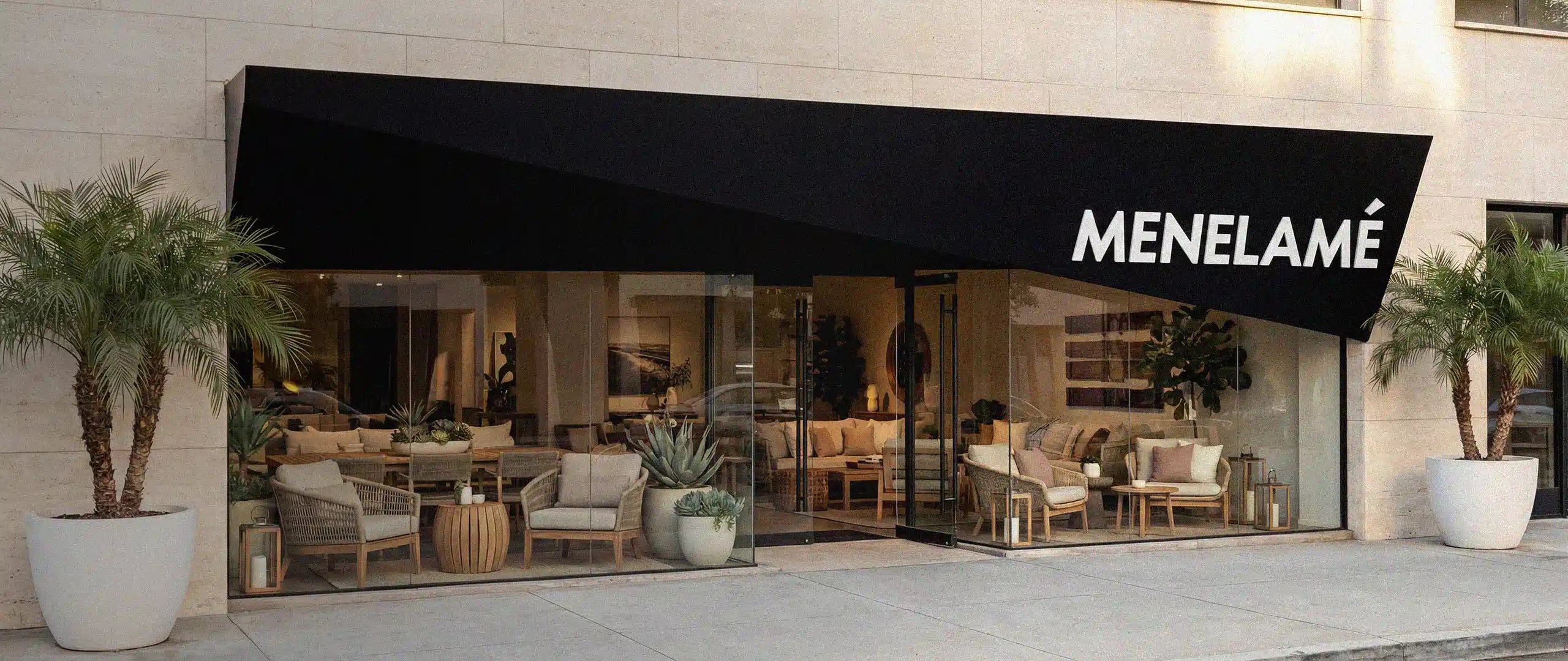 Außenansicht des modernen Menelamé Showrooms mit einer großen Glasfront und einer schwarzen Markise mit Markenschriftzug. Der Blick ins Innere zeigt exklusive Polstermöbel und Couchtische für hochwertiges Interieur-Design.