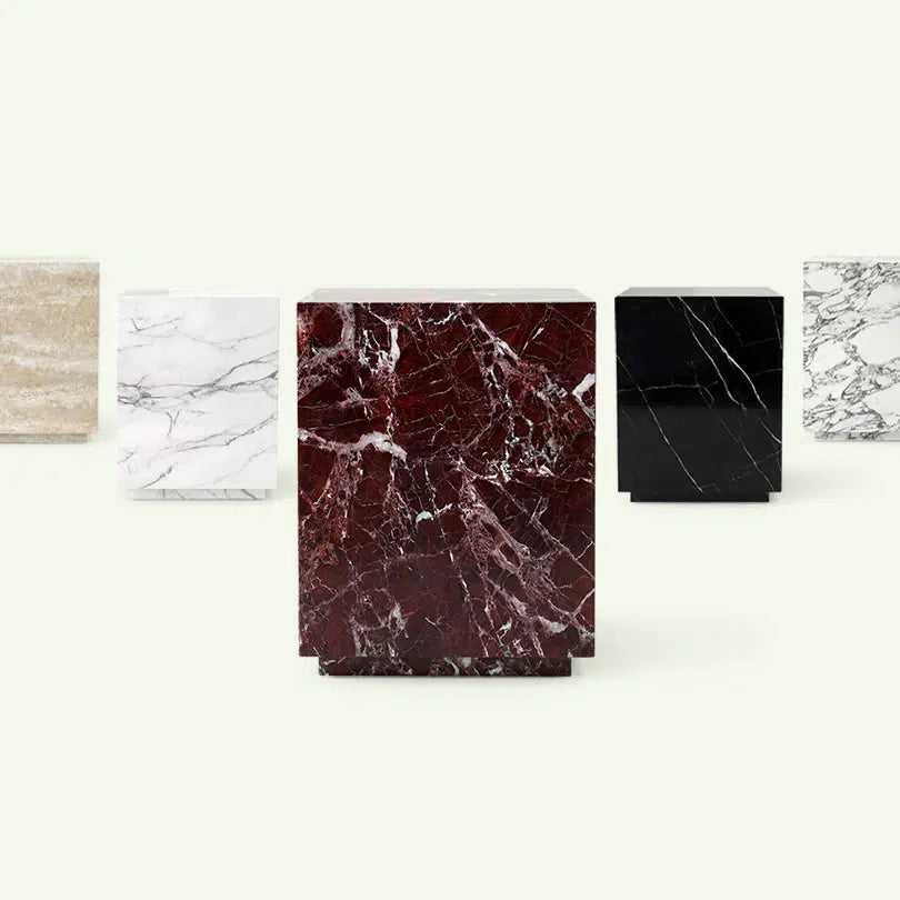 Materialübersicht der Pella Beistelltisch-Kollektion. Das Modell in dunkelrotem Rosso Levanto Marmor steht zentral im Fokus, umgeben von den Ausführungen in beigem Travertin, Calacatta White, Nero Marquina und Arabescato.