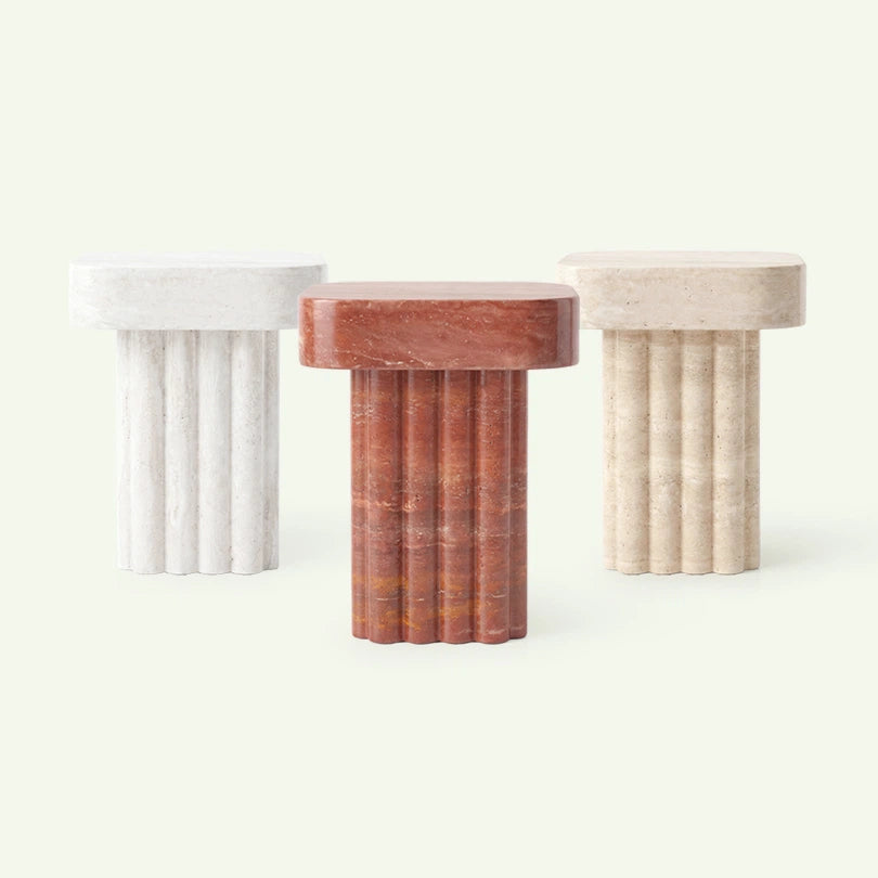Frontale Materialübersicht der Kutia Beistelltisch-Serie. Im Fokus steht das Modell aus rotem Travertin, flankiert von den Ausführungen in Super Weiß und klassischem beigem Travertin, um die architektonische Wirkung des Designs zu zeigen.