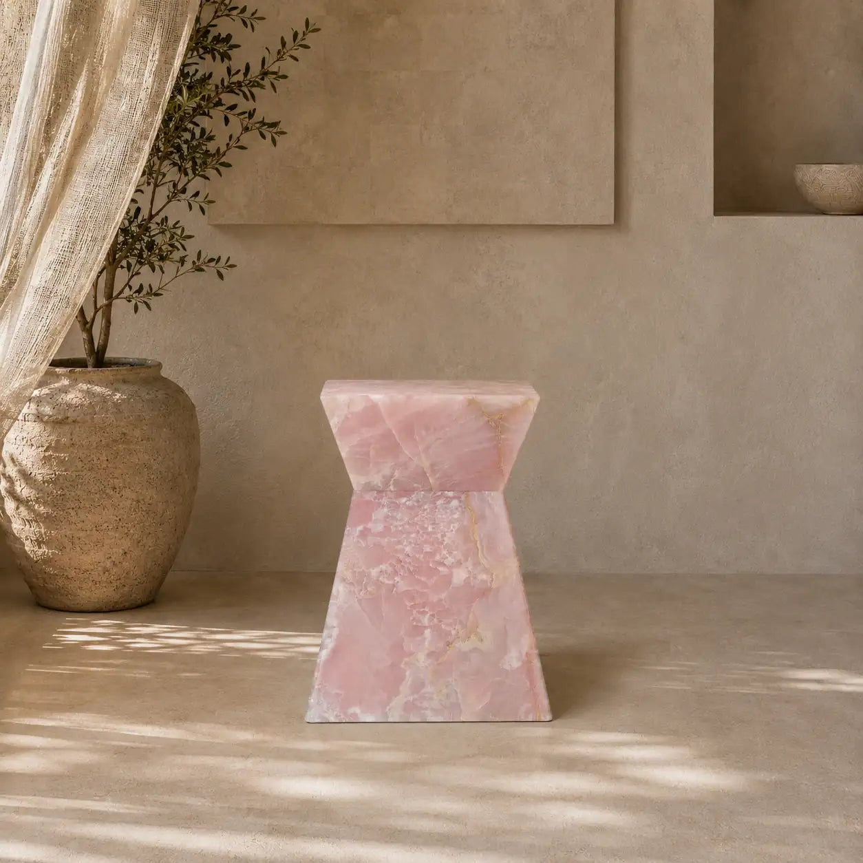Menelamé Shop by Look: Kunstvoller, monolithischer Beistelltisch in Sanduhr-Form aus seltenem, rosa gemasertem Naturstein "Pink Onyx" vor einer sanft ausgeleuchteten Wand.